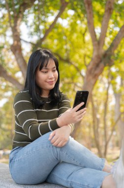 Yeşil bir parktaki bankta dinlenirken akıllı telefonunu kullanan genç bir Asyalı kadın. İnsanlar ve kablosuz teknoloji kavramları