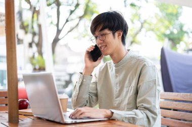 Asyalı bir erkek serbest meslek sahibi ya da iş adamı müşterisiyle telefonda konuşurken şehirdeki bir kafede çalışıyor..