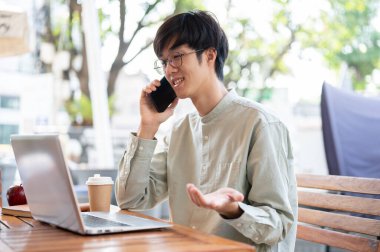 Asyalı bir erkek serbest meslek sahibi ya da iş adamı müşterisiyle telefonda konuşurken şehirdeki bir kafede çalışıyor..