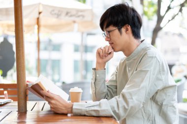 Parlak bir günde şehirde bir kafede dinlenirken kitap okumaya odaklanan yakışıklı bir Asyalı adamın yan görüntüsü..