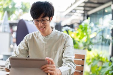 Mutlu bir Asyalı genç adam dijital tabletini kullanarak şehirdeki bir kahve dükkanında uzaktan çalışıyor. insanlar ve teknoloji kavramları