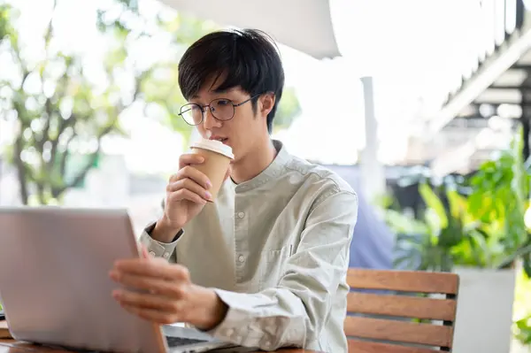 An Asian man is sipping coffee and focusing on his work on a laptop computer, working remotely at a cafe in the city. people and modern life concepts