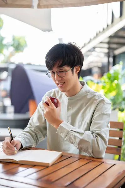 A handsome young Asian man is eating an apple while keeping his diary or writing down his ideas in a book at a cafe in the city.