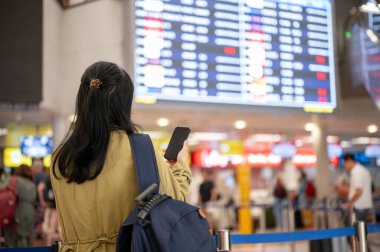 Asyalı bir kadın yolcunun ya da sırt çantasının arka plan görüntüsü havaalanındaki monitörden biniş saatini kontrol ediyor..