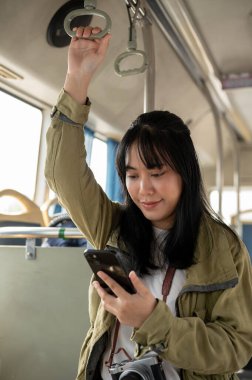 Umumi bir otobüste dururken akıllı telefonunu kullanan mutlu, genç bir Asyalı kadın yolcu. insanlar ve toplu taşıma kavramları