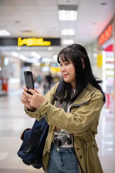 A happy young Asian female traveler or backpacker is using her smartphone while walking in the airport. boarding time, checking flight info, contacting someone