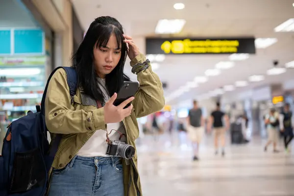 A confused young Asian female solo traveler is reading her flight information on her phone with a serious, doubtful face, facing a problem with self-check-in or missing her flight at the airport.