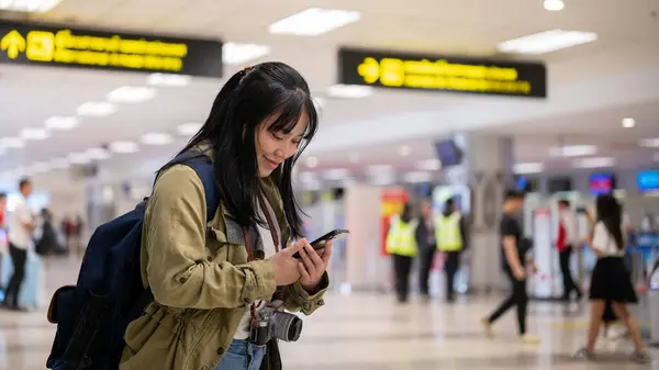 A positive young Asian female traveler is using her smartphone while standing in the airport. checking her flight info, booking taxi or hotel, doing self check-in online