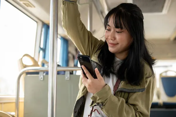 A happy young Asian female passenger using her smartphone while standing on a public bus. people and public transportation concepts
