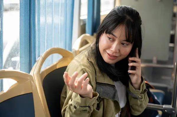 A smiling Asian female passenger is talking with someone on the phone while sitting in a bus, traveling by a public bus. public transportation and city life concepts