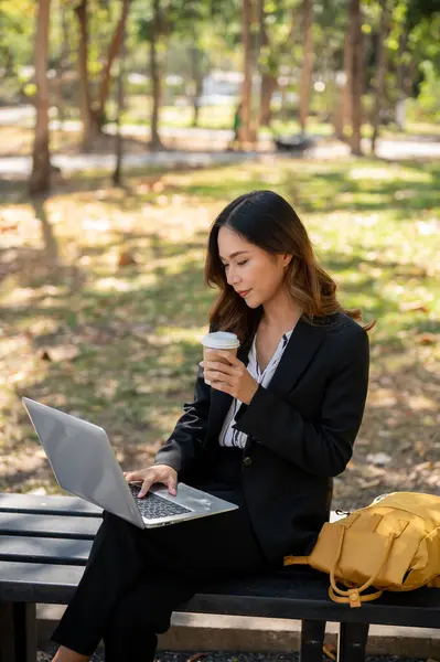 A beautiful Asian businesswoman is sipping coffee, sitting on a bench in a city park, and working on her laptop computer.