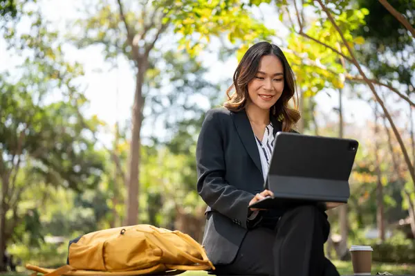 Parkta uzaktan çalışan çekici bir Asyalı iş kadını dijital tableti üzerinde çalışıyor..