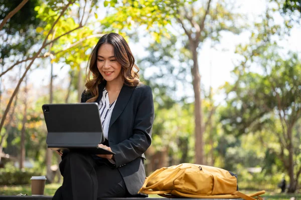Parkta uzaktan çalışan çekici bir Asyalı iş kadını dijital tableti üzerinde çalışıyor..