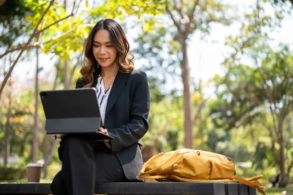 Parkta uzaktan çalışan çekici bir Asyalı iş kadını dijital tableti üzerinde çalışıyor..