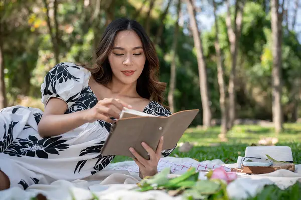 A gorgeous, positive Asian woman in a cute dress is reading a book on a picnic mat while picnicking in a green park on a bright day. leisure and hobby concepts