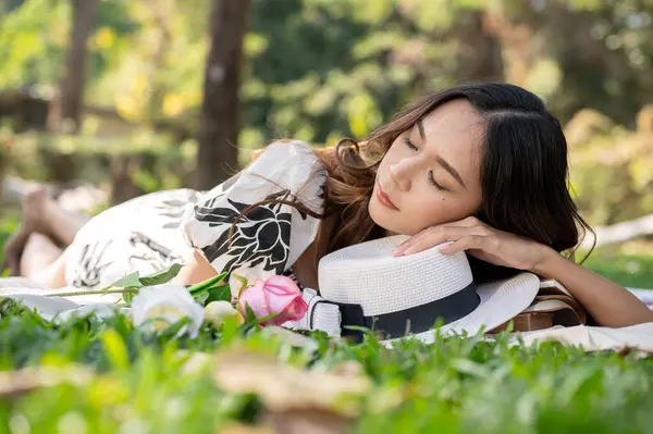 A carefree and attractive Asian woman is falling asleep or taking a nap on a picnic mat in a green park or garden.