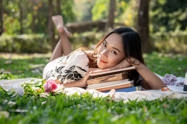 An attractive Asian woman is lying on a picnic mat, putting her head on a stack of books, and enjoying a picnic in a green garden or park on a bright day.