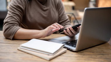 Şehirdeki bir kafede uzaktan çalışan, akıllı telefonunu kullanarak dizüstü bilgisayarının önünde oturan bir kadının görüntüsü..
