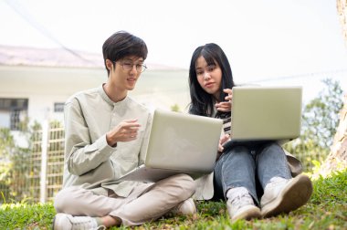 İki Asyalı üniversite öğrencisi, kampüs parkında çimlerin üzerinde otururken bilgisayar ekranına bakarak, konuşarak ve fikirlerini paylaşarak iş birliği yapıyorlar..