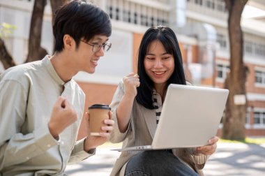 İki neşeli Asyalı üniversite öğrencisi yumruklarını gösteriyor, dizüstü bilgisayara bakıyor ve üniversite parkında otururken birlikte sınav sonuçlarını kutluyorlar..