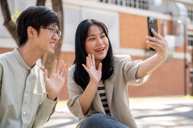 İki neşeli Asyalı arkadaş, yemek molasında şehir parkında dinlenirken akıllı telefonlarıyla fotoğraf çekiyorlar..