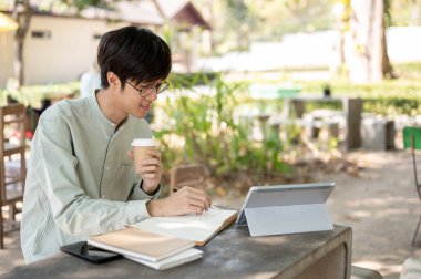 Olumlu ve zeki Asyalı bir adam bir kafede uzaktan çalışıyor, açık bir masada oturuyor, online çalışıyor, ya da ödev yapıyor..