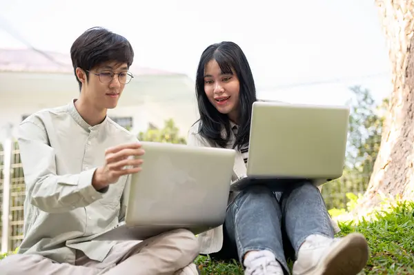 Two Asian university students are collaborating on work, looking at the laptop screen, talking, and sharing their ideas on a project while sitting on the grass in the campus park.