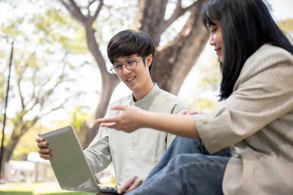A happy Asian male university student is using his laptop computer and working with his university friend in the park on a bright day.
