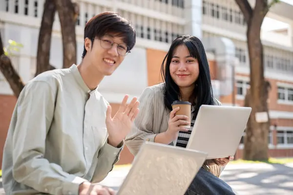 Two smiling Asian university students are sitting on a bench in the university park together with their laptop computer, smiling at the camera.
