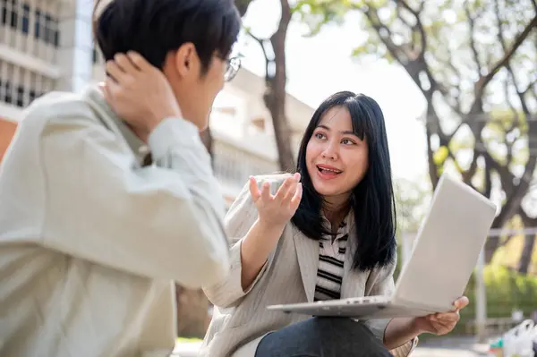 Two smart Asian university students are discussing and collaborating on a project while sitting together on a bench in the university park. university life, friendship, studying