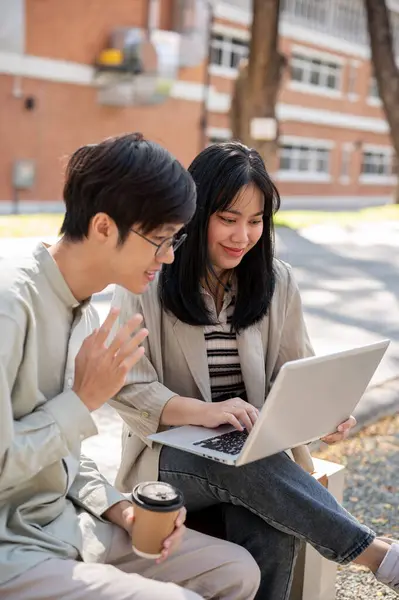Two smart Asian university students are discussing and collaborating on a project while sitting together on a bench in the university park. university life, friendship, studying