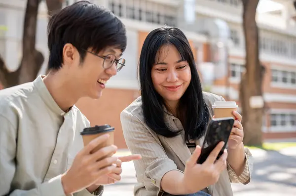 Two cheerful Asian friends are enjoying talking, watching videos, or reading online articles on a smartphone together while chilling in a city park on their lunch break.