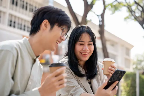 Two cheerful Asian friends are enjoying talking, watching videos, or reading online articles on a smartphone together while chilling in a city park on their lunch break.