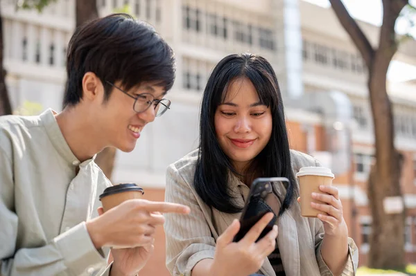 两个快乐的亚洲朋友正在享受聊天 看视频或一起用智能手机阅读网上文章的乐趣 而在他们午休的时候 他们则在城市公园里寒战一番 — 图库照片
