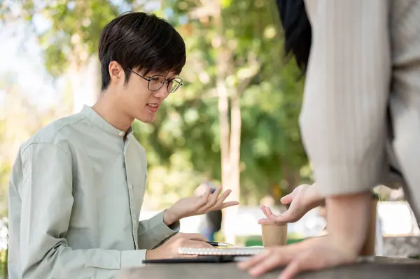 A smart Asian male university student is explaining and sharing his ideas with his friend while collaborating on work together in the university park.
