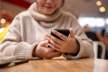 Rahat bir kazak giymiş bir kadının kafedeki bir masada akıllı telefonunu kullanırken görüntüsü. Sohbet ediyor, mesajlara cevap veriyor ve bir web sitesine kayıyor..