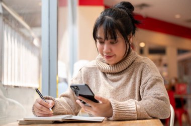 Rahat örülü süveter giyen pozitif bir Asyalı kadın, çok yönlü iş yapıyor, defterine not alıyor, akıllı telefonuyla bazı bilgiler okuyor ve bir kafede uzaktan çalışıyor..