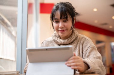 Rahat örgü kazaklı, güzel, pozitif Asyalı bir kadın dijital tabletini kullanıyor ve bir kafede uzaktan çalışıyor. İnsanlar ve kablosuz teknoloji kavramları