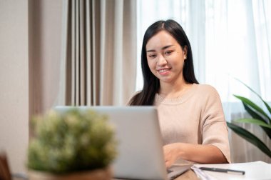 Güzel ve pozitif Asyalı bir kadın ev ofisinin çalışma alanında dizüstü bilgisayarı üzerinde çalışıyor, evde uzaktan çalışıyor..