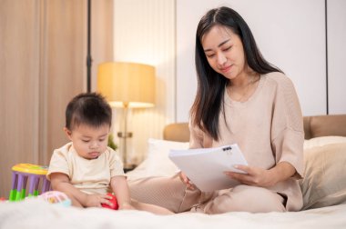 Mutlu, sevimli bir çocuk oyuncaklarla oynarken annesi yatakta çalışıyor, evrak okuyor ve yatak odasında birlikte vakit geçiriyor..
