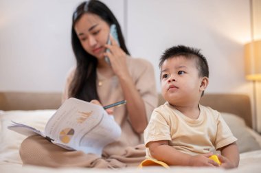 Sevimli, masum bir Asyalı çocuk yatakta oyuncak oynarken annesi çalışıyor ve telefonda konuşuyor. çocukluk ve annelik kavramları