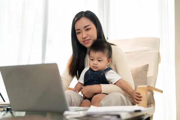 A beautiful Asian businesswoman mom is working remotely at home, focusing on her laptop computer while taking care of her little son. Work from home and mom\'s life concepts.