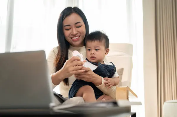 A busy Asian working woman mom is multitasking, working remotely from home, focusing on her work and taking care of her cute little son in the living room. mom\'s life concept