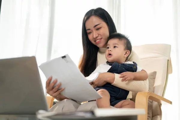 A busy Asian working woman mom is multitasking, working remotely from home, focusing on her work and taking care of her cute little son in the living room. mom\'s life concept