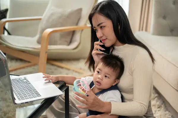 A positive Asian businesswoman mom is talking on the phone with her colleague while working from home and taking care of her little son. working remotely and mom\'s life concepts