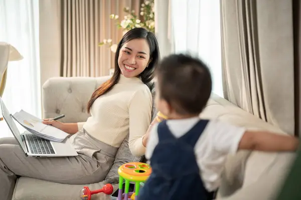 A happy Asian businesswoman mom is talking and playing with her baby boy on a sofa in the living room while working from home. busy mom\'s life, family bonding, single mom