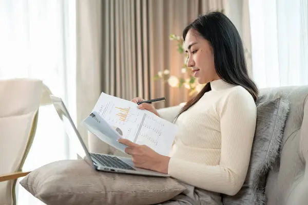 A beautiful Asian businesswoman is working remotely from home, reading documents, and focusing on her laptop computer.