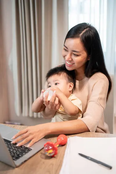 A positive Asian mom is feeding milk from a bottle to her baby boy while working on her laptop computer in the living room. work from home and mom\'s life concepts