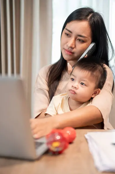 A busy Asian businesswoman mom is multitasking, talking on the phone with her client, focusing on her laptop, and taking care of her little son at home. work from home and mom\'s life concepts