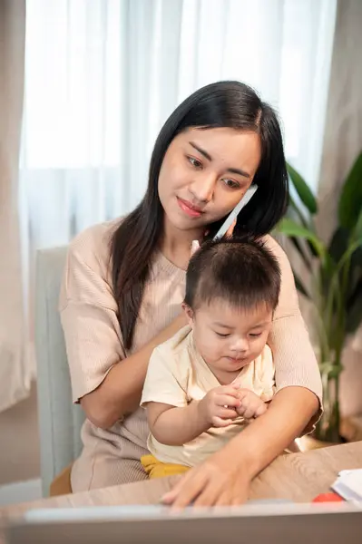 A busy Asian businesswoman mom is multitasking, talking on the phone with her client, focusing on her laptop, and taking care of her little son at home. work from home and mom\'s life concepts